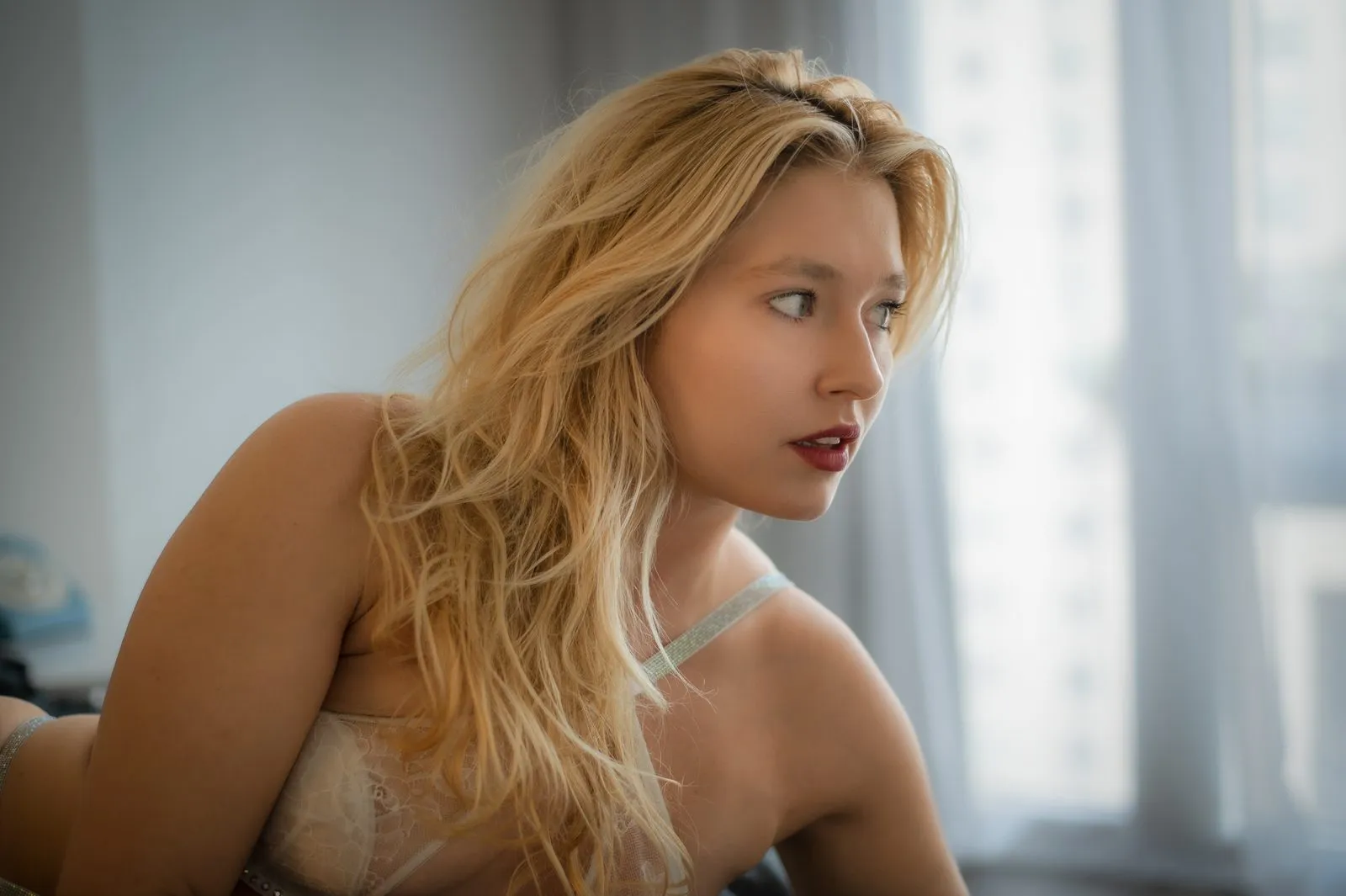 A close-up profile portrait of a blonde woman in lace lingerie, gazing out a window with a soft Manhattan skyline visible in the background.